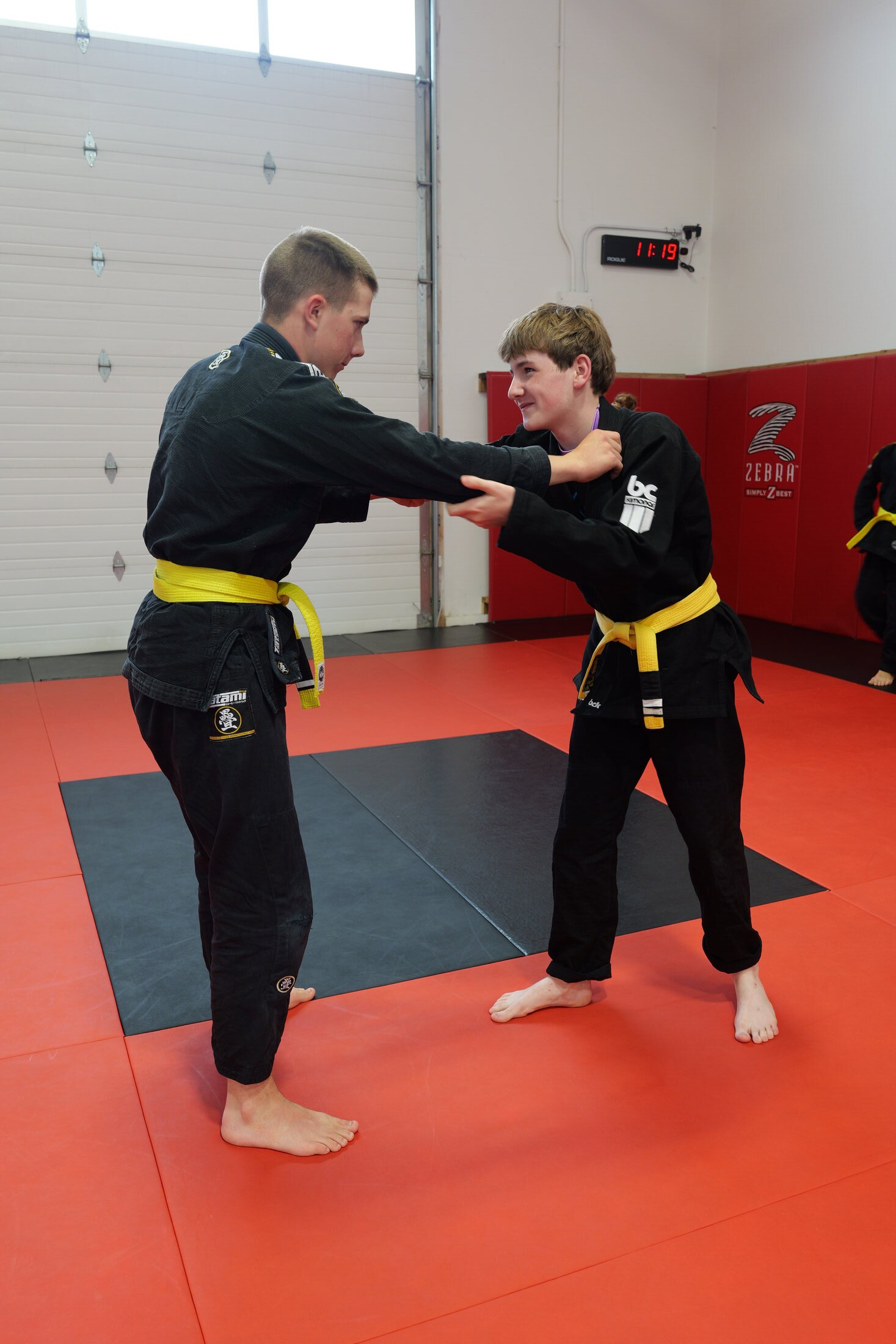 Two teenage Brazilian Jiu Jitsu students in black gis and yellow belts grip fighting from standing on the red mats of the Arashi-Do Red Deer training floor