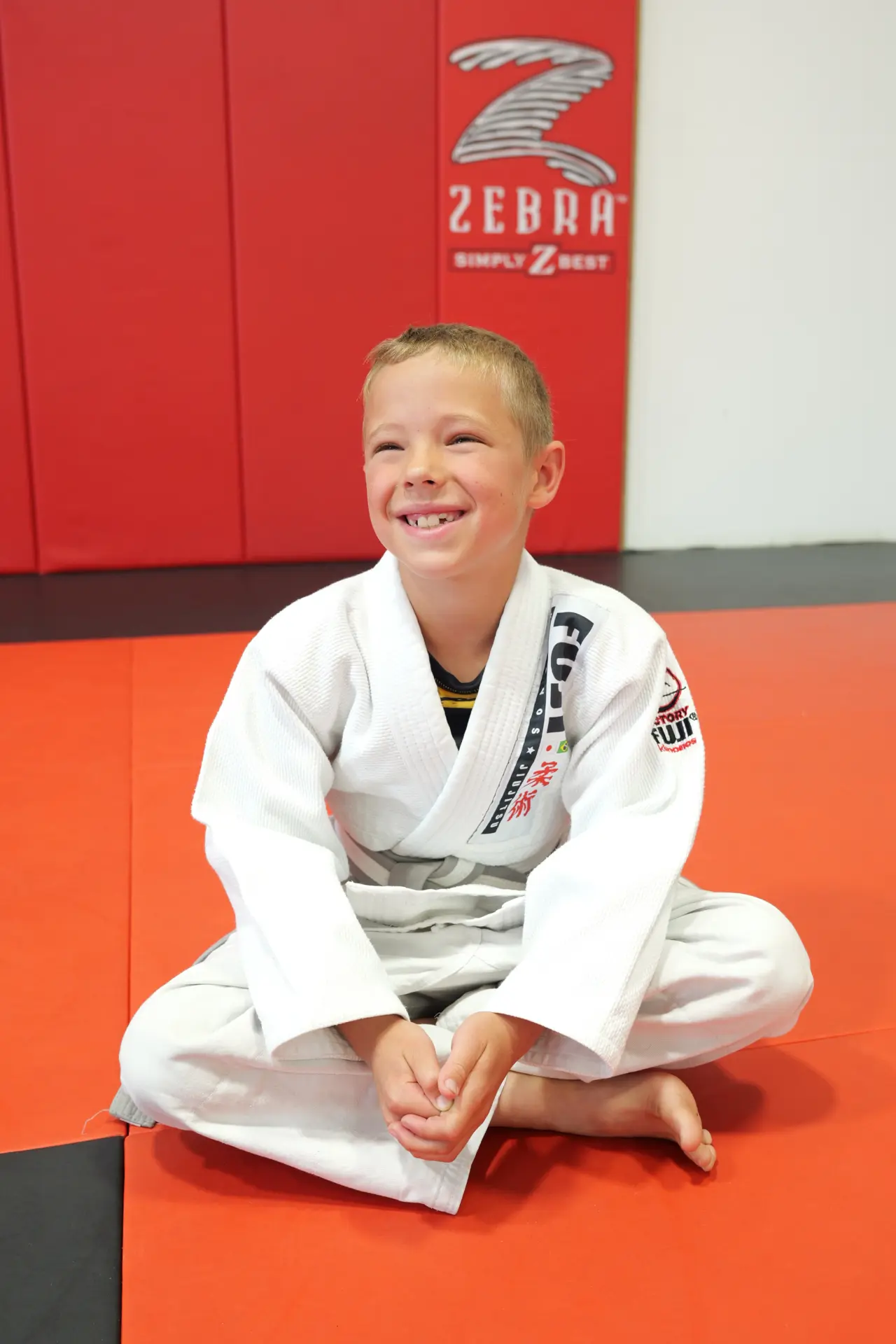 Young white-belt student in a white gi smiling while sitting on the red and grey mats at Arashi-Do Red Deer